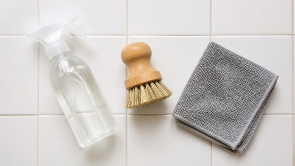 A spray bottle of white vinegar, a scrubbing brush and a microfibre cloth on a tiled bathroom surface, seen from above