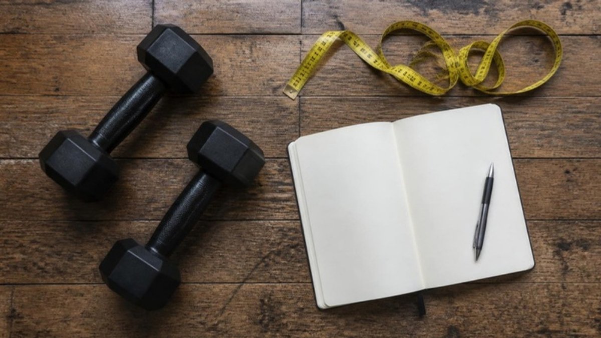 Two dumbbells, a tape measure and an open notebook with a pen on a wooden floor, seen from above