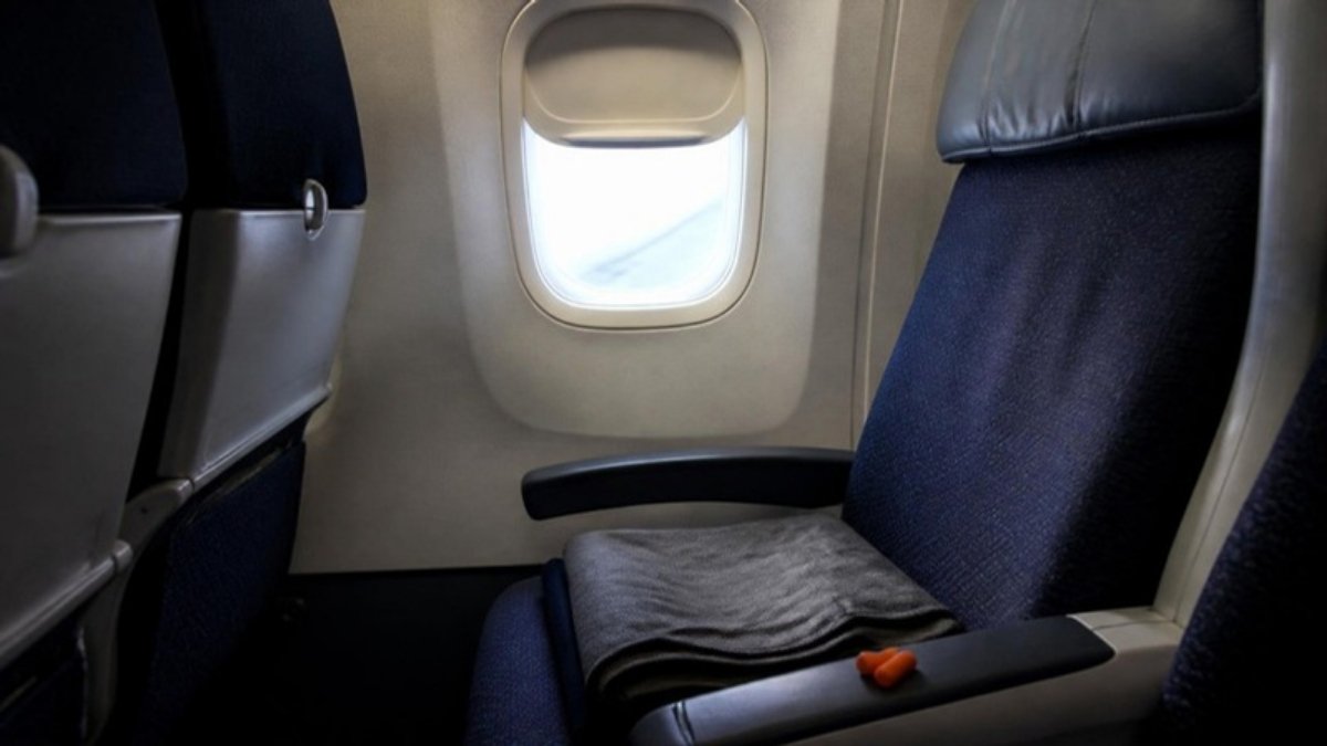 An airplane window seat with the shade half down, a grey blanket folded on the seat and a pair of earplugs on the armrest