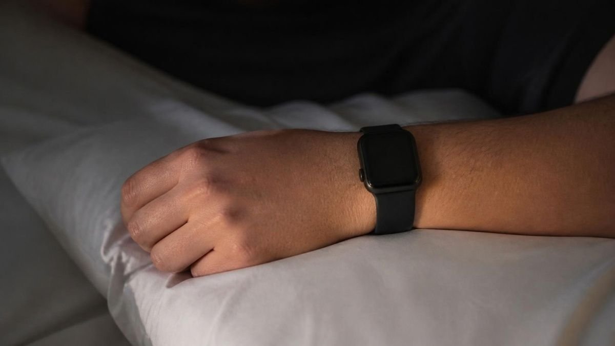 The hand and wrist of a sleeping person wearing a black smartwatch, resting on a white pillow in dim light