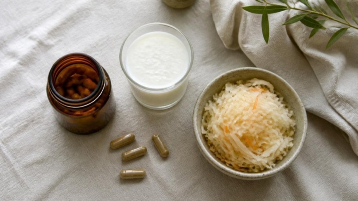 A jar of probiotic capsules with a few spilled out beside a glass of kefir and a bowl of sauerkraut, seen from above