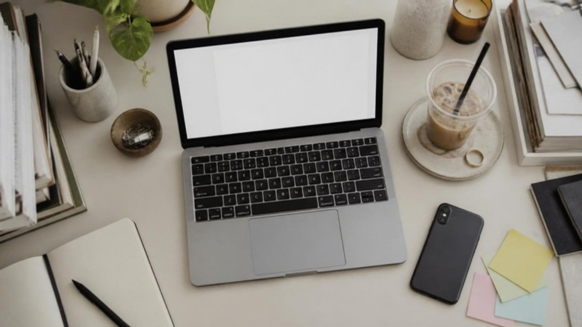 An open laptop with a blank document, a phone face down beside it and a cold cup of coffee on a cluttered desk, seen from above