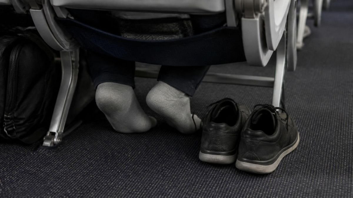 A passenger’s feet in socks resting under an economy seat on a long-haul flight, shoes loosened beside them on the cabin carpet